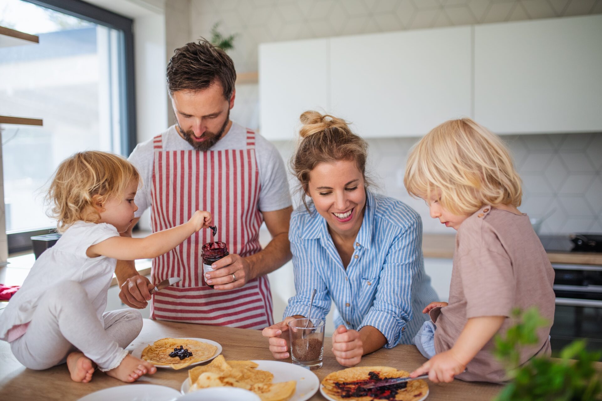 Young family with two small children indoors in kitchen, eating pancakes.