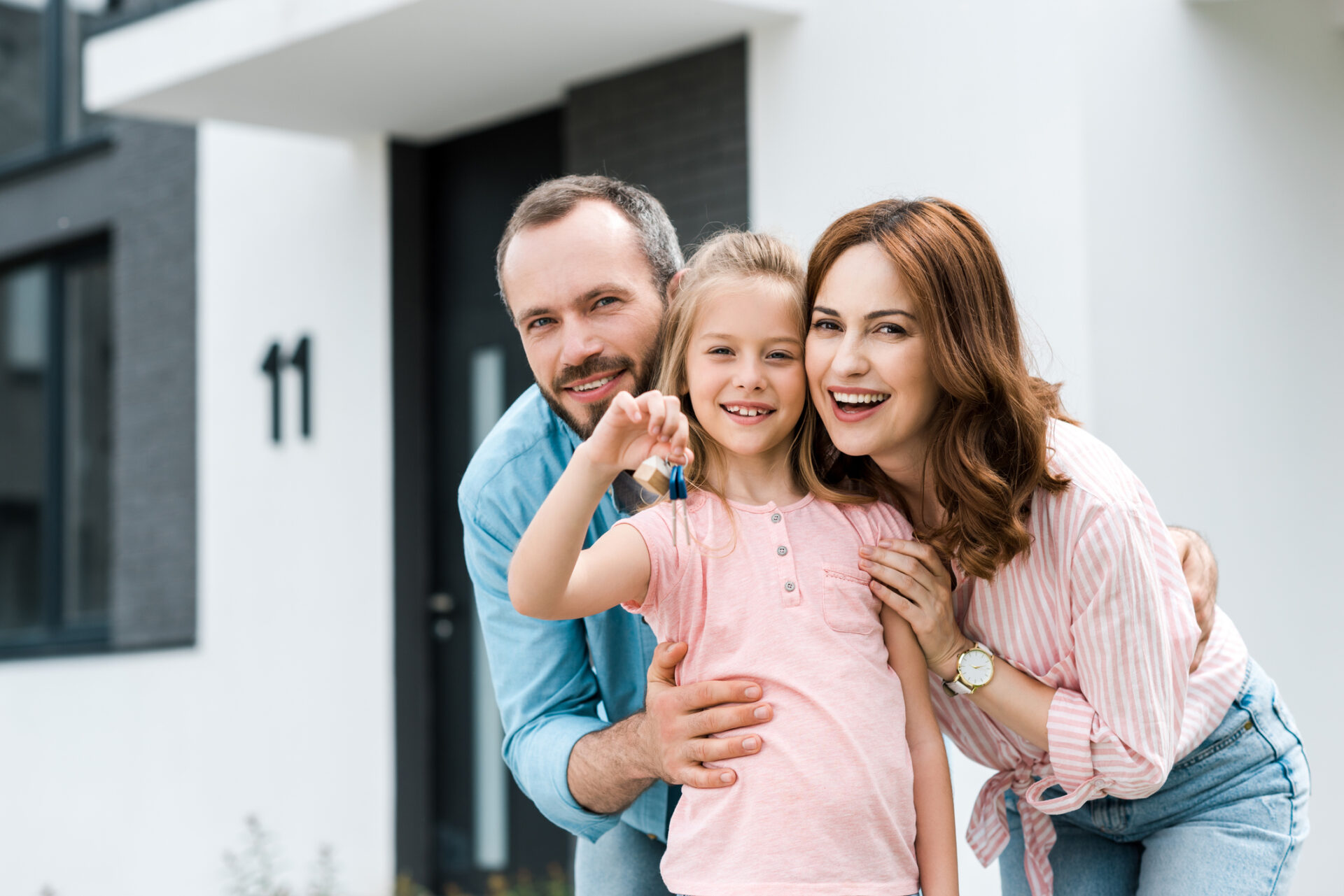 happy kid holding key near mother and father while standing near house and sign with sold letters