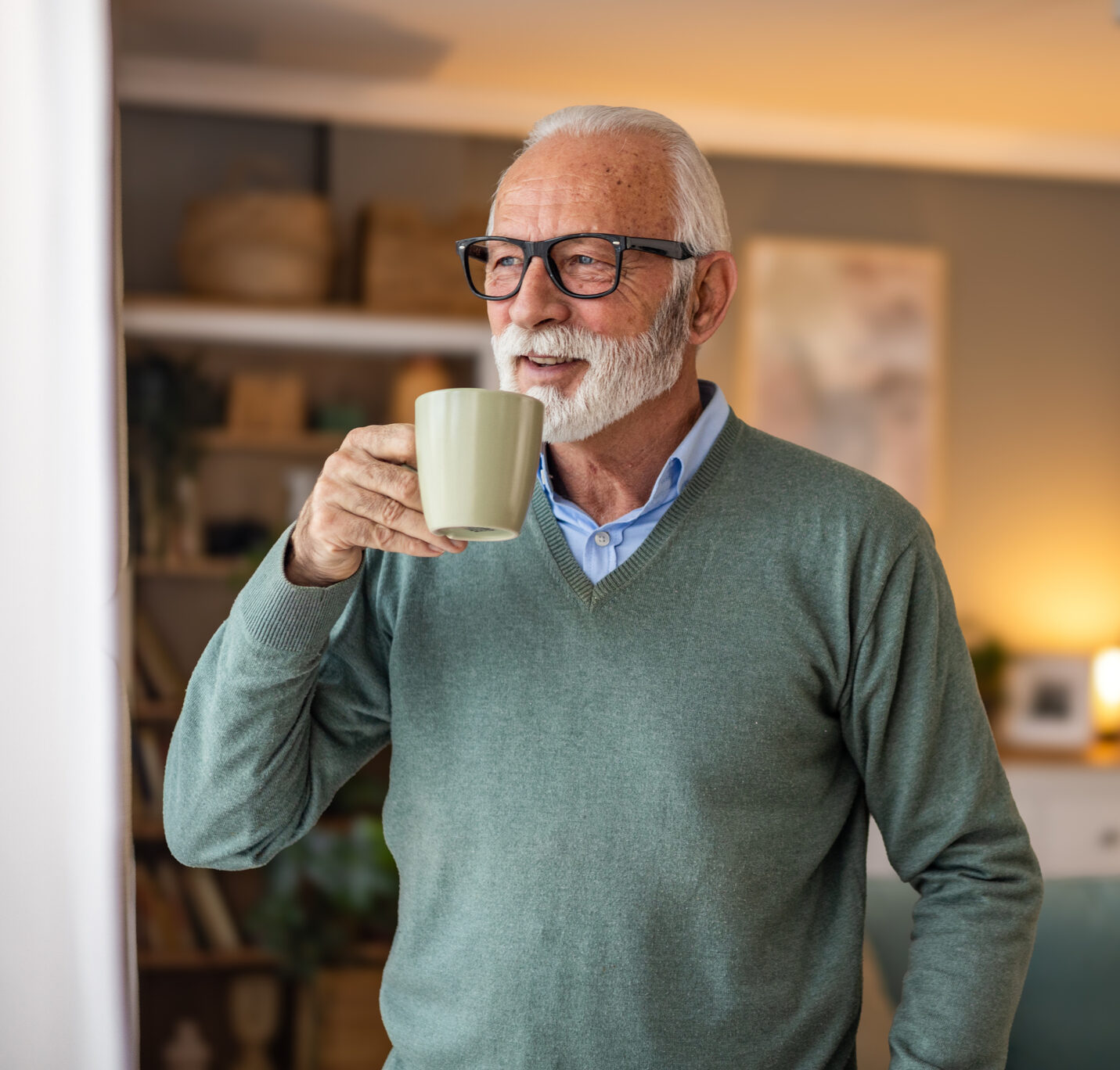 An elderly man is standing in the living room of a nursing home with a happy smile and holding a cup of coffee